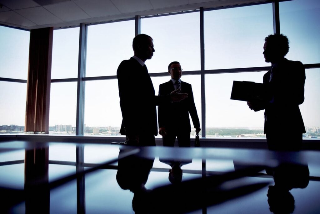 The silhouettes of three law firm partners engaged in a high-stakes strategic meeting in a modern, high-rise conference room. This represents the top-level focus that firm leadership can achieve once their client intake and sales systems are optimized by the Firm Heartbeat experience.