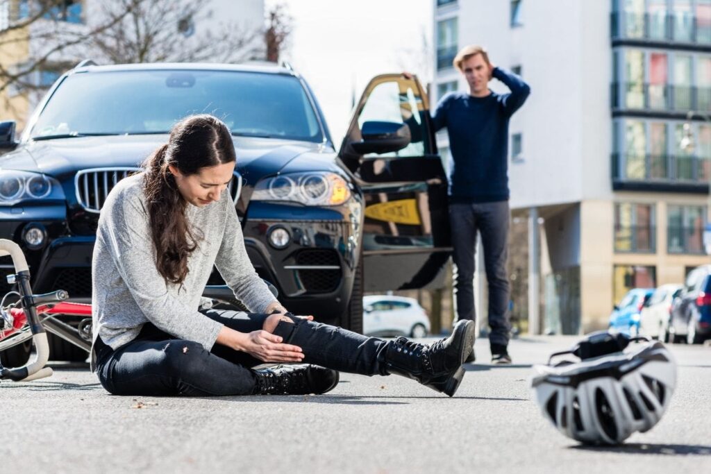 A young woman with long dark hair sits on the pavement of a street after a bicycle accident, cradling her injured knee with both hands and looking down in pain. Her bicycle lies on its side next to her, and her helmet is on the ground in the foreground. In the background, a concerned man stands by the open driver's side door of a black SUV, with his hand on his head in a distressed gesture.