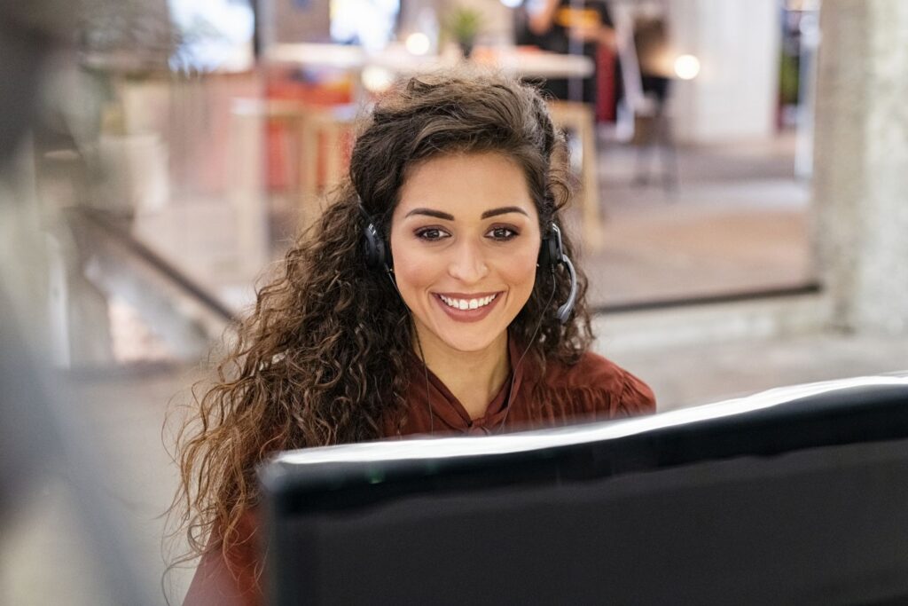 A confident and engaging female intake specialist for a law firm, smiling directly at the camera from her desk in a modern office. She wears a headset, embodying the skilled, client-focused sales professional developed through the Firm Heartbeat experience.