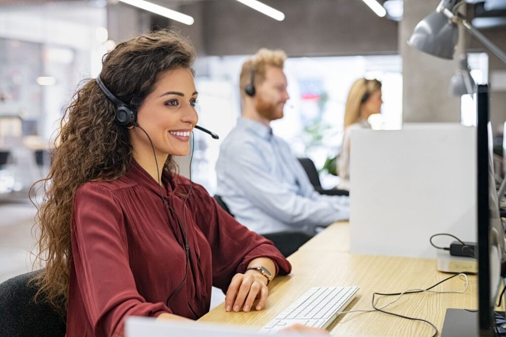 A vibrant image of a professional, diverse law firm intake or call center team. In the foreground, a smiling woman with long curly hair wears a headset and looks at her computer screen, embodying excellent client service. Two other team members are visible in the background, also at their workstations with headsets, symbolizing an efficient and well-trained client acquisition operation, a core benefit of the Firm Heartbeat experience.