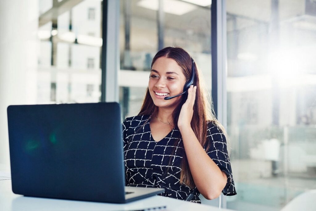 A highly professional female intake specialist working in a bright, modern law office with large windows. She is smiling warmly while engaging with a client on her headset, representing the clarity, positivity, and success that the Firm Heartbeat experience brings to a firm's sales and intake process.