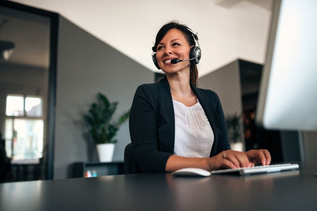 A skilled female intake specialist for a law firm, actively listening and smiling while engaging with a potential client on her headset in a modern office. Her professional demeanor and readiness to capture information exemplify the expert communication skills developed through the Firm Heartbeat experience.
