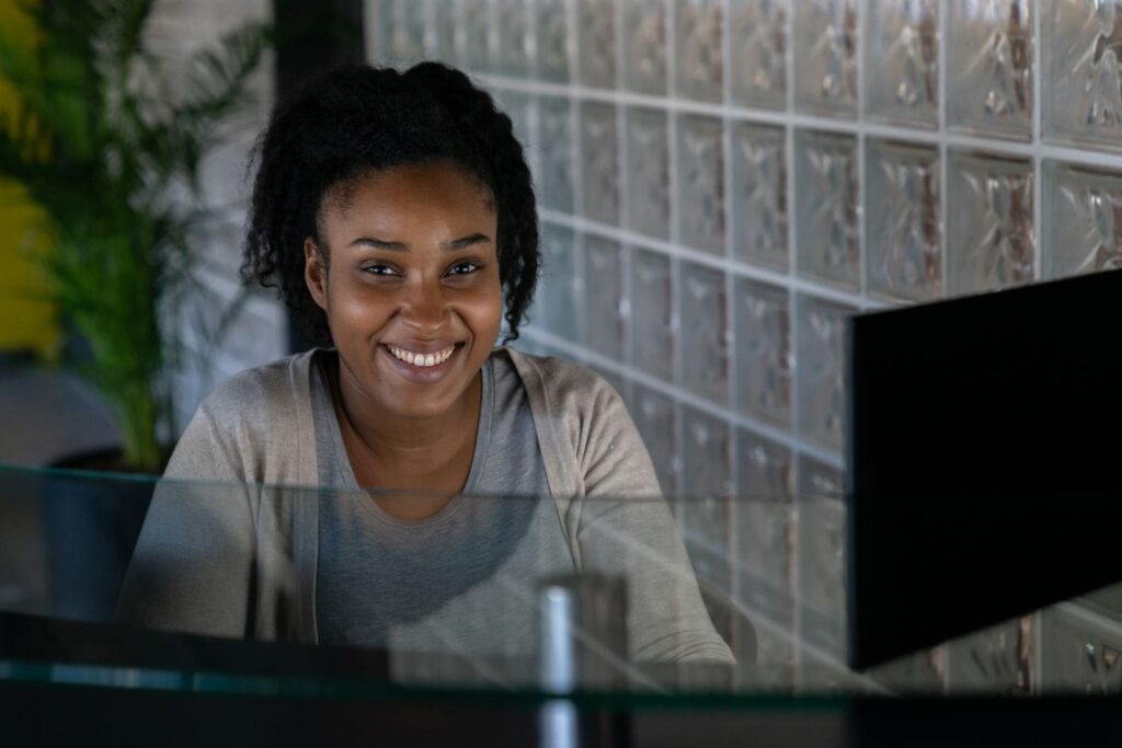 A welcoming Black female receptionist for a law firm, smiling warmly and directly at the camera from her desk. Her genuine, empathetic expression represents the trust and human connection that the Firm Heartbeat experience builds into your client intake process.