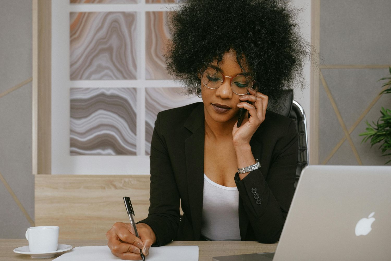A professional Black female sales specialist for a law firm, intently listening on a phone call while taking meticulous notes at her modern desk. Her focused execution represents the elite skill set and disciplined process installed by the Firm Heartbeat experience to maximize client conversion.