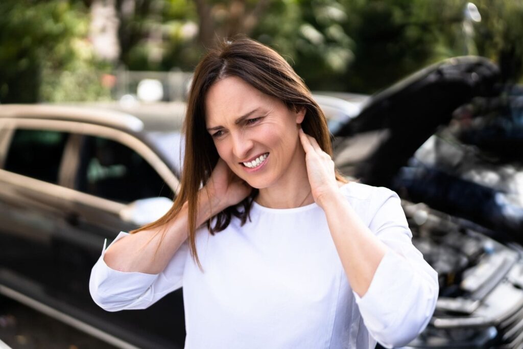 A middle-aged woman with shoulder-length brown hair stands outdoors, grimacing in pain as she clutches the back of her neck with both hands. The background is blurred but shows the front of a dark-colored car with its hood open, suggesting she has just been in a car accident.