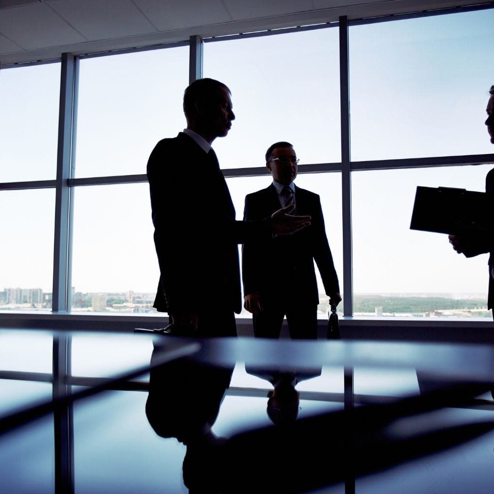 The silhouettes of three law firm partners engaged in a high-stakes strategic meeting in a modern, high-rise conference room. This represents the top-level focus that firm leadership can achieve once their client intake and sales systems are optimized by the Firm Heartbeat experience.