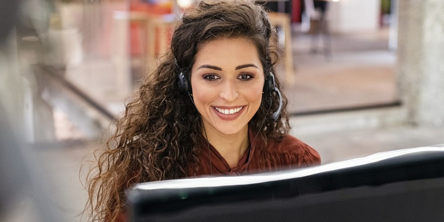 A confident and engaging female intake specialist for a law firm, smiling directly at the camera from her desk in a modern office. She wears a headset, embodying the skilled, client-focused sales professional developed through the Firm Heartbeat experience.