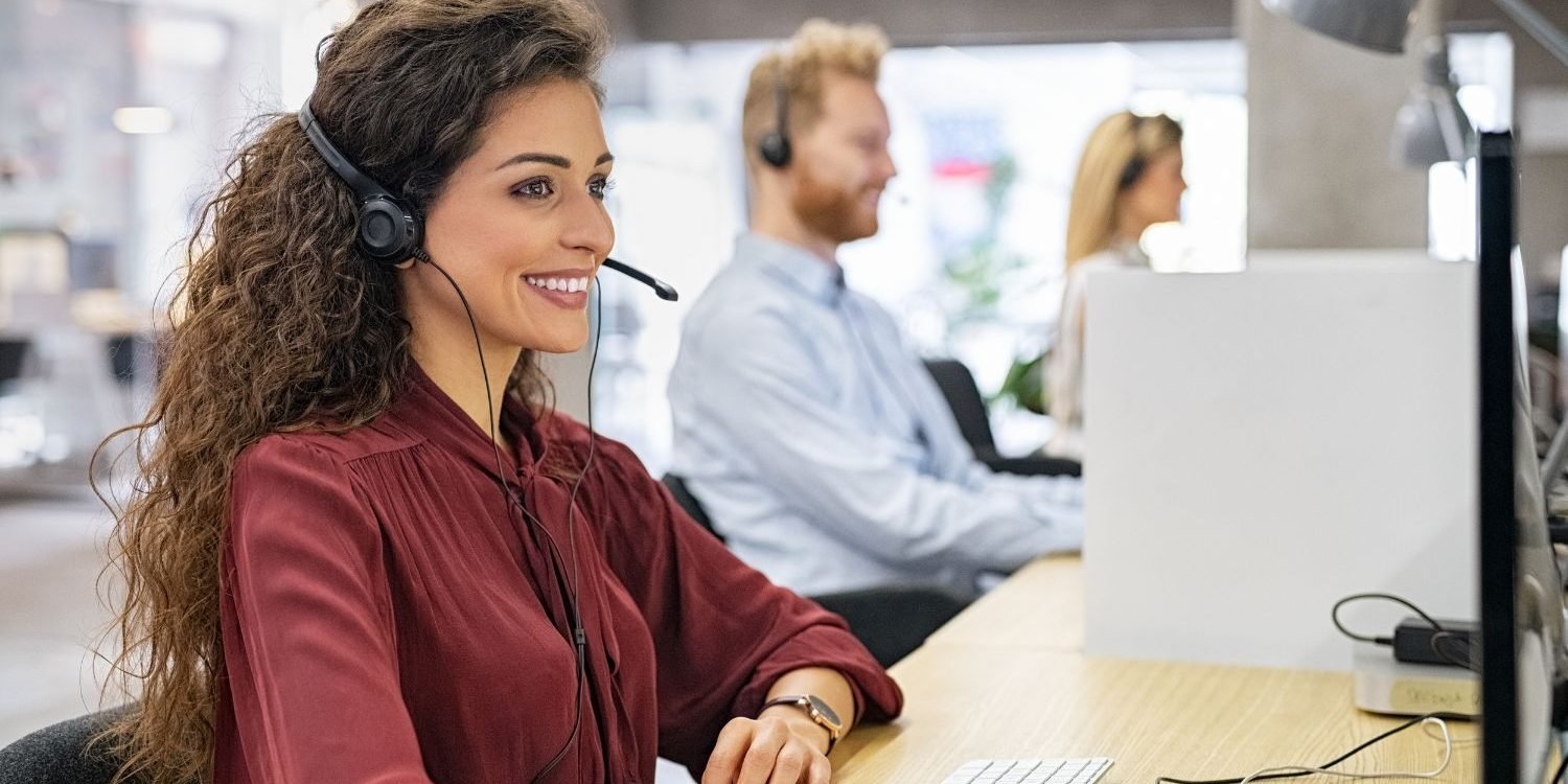 A vibrant image of a professional, diverse law firm intake or call center team. In the foreground, a smiling woman with long curly hair wears a headset and looks at her computer screen, embodying excellent client service. Two other team members are visible in the background, also at their workstations with headsets, symbolizing an efficient and well-trained client acquisition operation, a core benefit of the Firm Heartbeat experience.