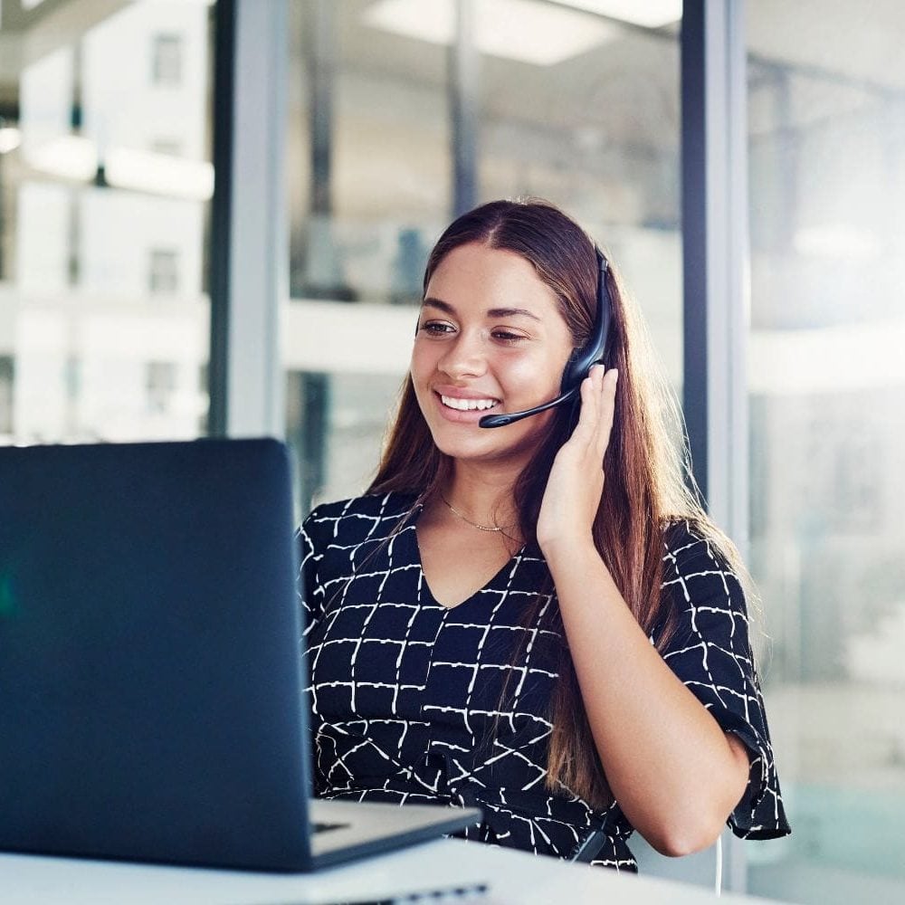 A highly professional female intake specialist working in a bright, modern law office with large windows. She is smiling warmly while engaging with a client on her headset, representing the clarity, positivity, and success that the Firm Heartbeat experience brings to a firm's sales and intake process.