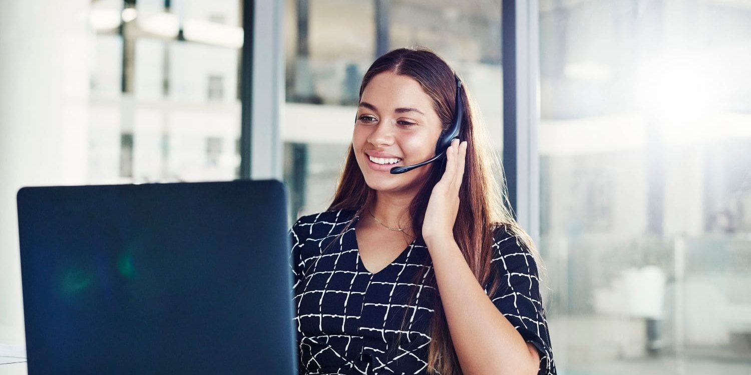 A highly professional female intake specialist working in a bright, modern law office with large windows. She is smiling warmly while engaging with a client on her headset, representing the clarity, positivity, and success that the Firm Heartbeat experience brings to a firm's sales and intake process.