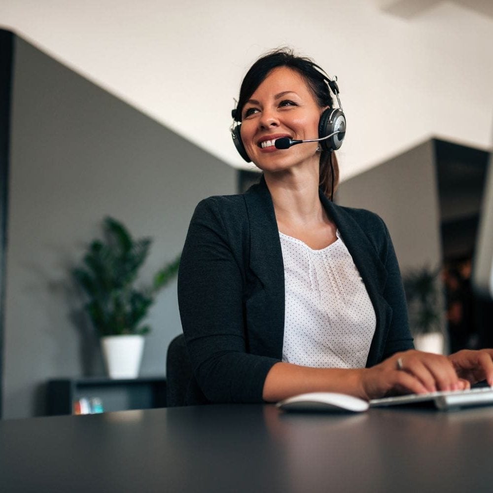A skilled female intake specialist for a law firm, actively listening and smiling while engaging with a potential client on her headset in a modern office. Her professional demeanor and readiness to capture information exemplify the expert communication skills developed through the Firm Heartbeat experience.