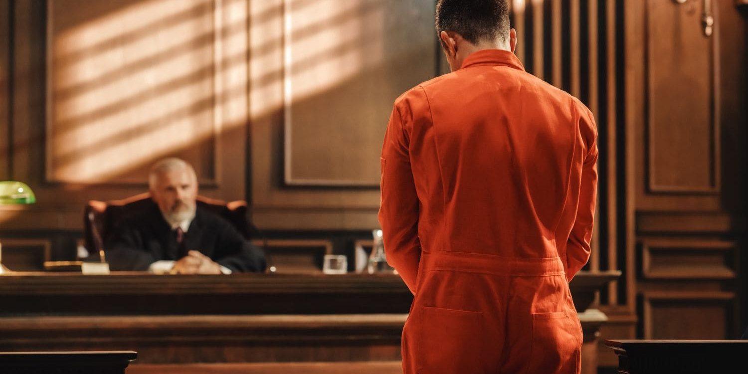 A defendant in an orange jumpsuit stands before a judge in a solemn courtroom, their back to the viewer, symbolizing the high-stakes nature of criminal defense cases. This dramatic scene highlights the critical importance of a law firm's sales and intake process in securing representation for individuals facing such pivotal moments, a process optimized by the Firm Heartbeat experience.