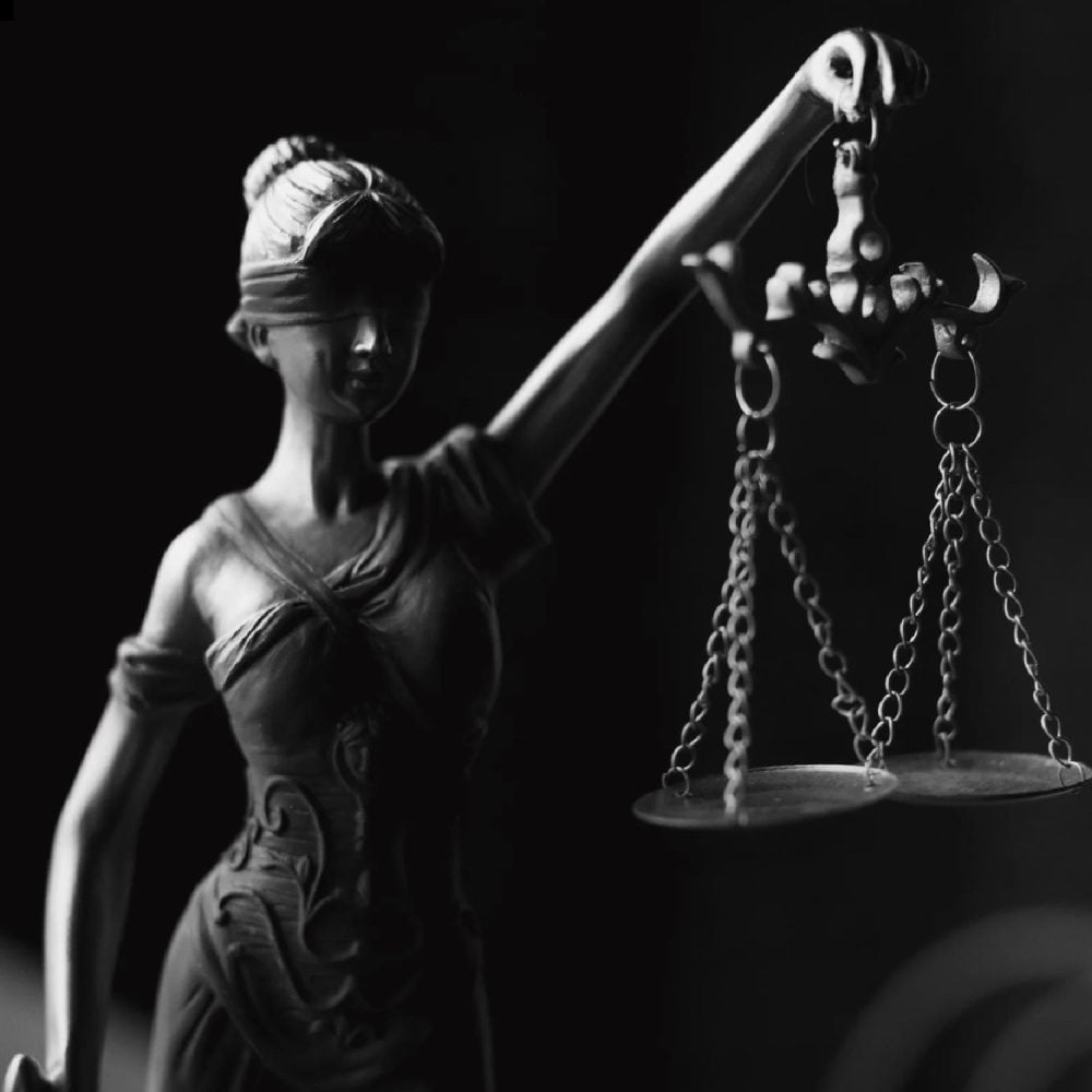 A dramatic, high-contrast, black and white photograph of the Lady Justice statue holding her iconic scales. The image symbolizes the need to balance the principled practice of law with the profitable business of law, a core mission of the Firm Heartbeat experience for law firms.
