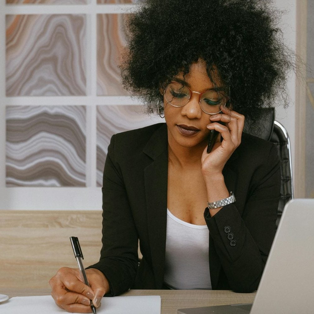 A professional Black female sales specialist for a law firm, intently listening on a phone call while taking meticulous notes at her modern desk. Her focused execution represents the elite skill set and disciplined process installed by the Firm Heartbeat experience to maximize client conversion.