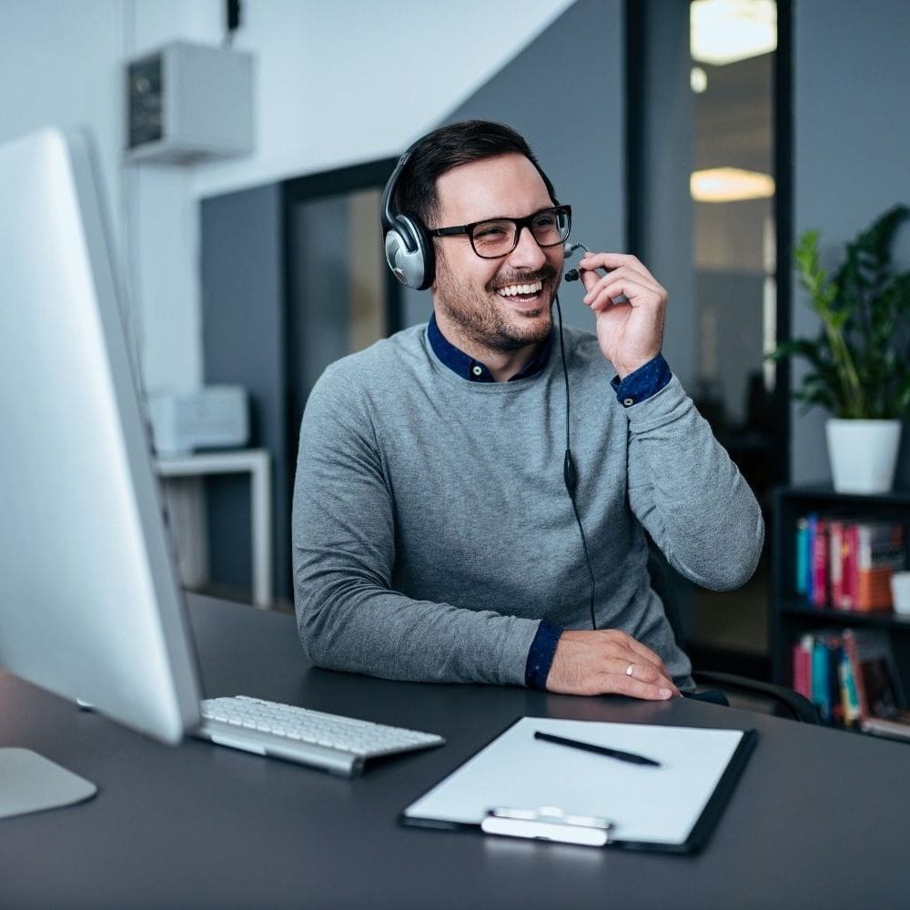 A male intake specialist with dark hair, glasses, and a headset laughs warmly while speaking to someone on a call in a modern office. He is seated at his desk in front of a computer, holding the microphone piece of his headset, creating a positive and engaging customer service image.