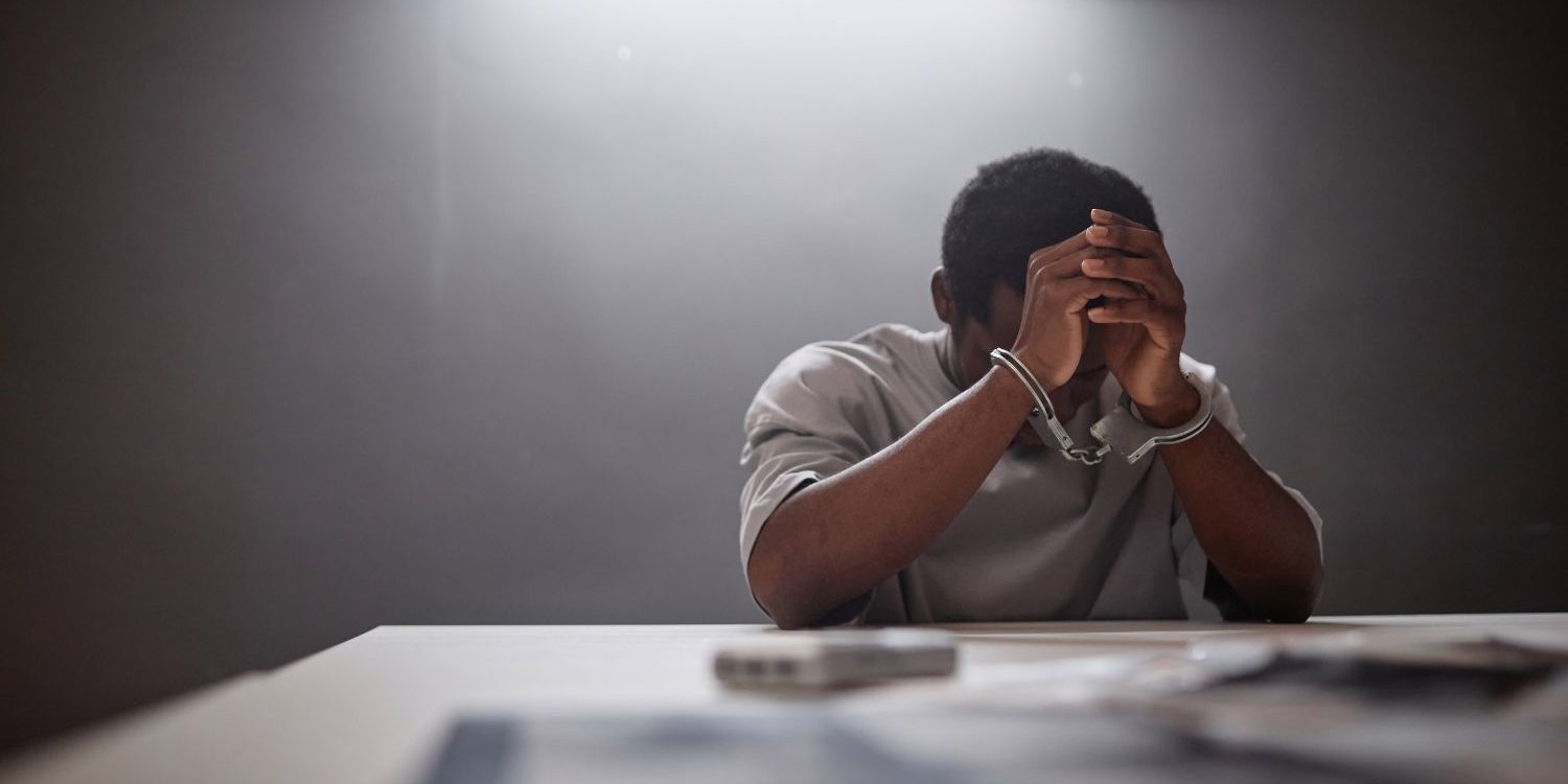 A dramatic and somber photograph of a man in an interrogation room. A young Black man, wearing a plain, light-gray t-shirt, sits at a light-colored table with his head bowed and his face buried in his hands in a gesture of deep distress, shame, or despair. Metal handcuffs are clearly visible, binding his wrists together. The room is dark and stark. A single, harsh fluorescent light bar on the ceiling casts a focused, downward beam, intensely illuminating the man and the table while the background fades into deep shadow. In the blurred foreground on the table, there are some indistinct items, possibly case files or evidence. The image's high-contrast lighting and the man's body language create a powerful atmosphere of isolation, regret, and confinement.