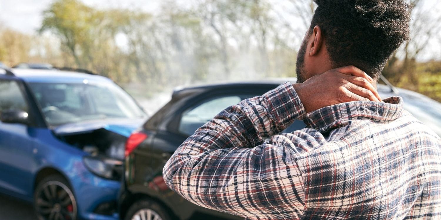 An over-the-shoulder shot of a man standing at the scene of a car accident, seen from his back and right side. He is a Black man with short, textured hair, wearing a plaid button-down shirt. He is holding the back of his neck with his left hand, a gesture that suggests pain or a whiplash injury from the collision. In the blurred background in front of him, at least two cars are damaged. On the left, a metallic blue car has significant front-end damage; its hood is crumpled and popped open, with faint steam or smoke rising from the engine bay. Next to it is a dark-colored car. The setting is outdoors on a road, with out-of-focus trees under a bright, overcast sky. The focus on the man highlights the human element and potential injury resulting from the accident.