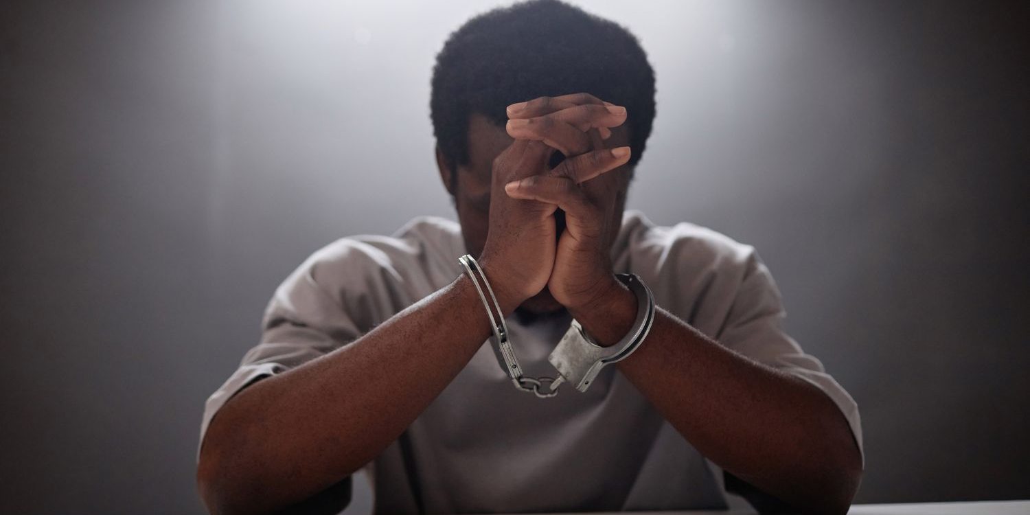 A dramatic, dimly lit photo of a Black man sitting at a table in what appears to be an interrogation room. He is wearing handcuffs, and his head is bowed as he covers his face with his clasped hands, conveying a strong sense of stress, despair, or contemplation.
