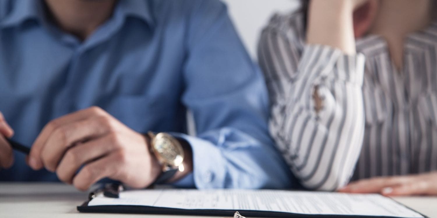 A poignant, close-up photograph symbolizing divorce or marital separation. The sharp focus is on a gold wedding band and a silver engagement ring lying on a white table in the foreground, placed in front of a document on a black clipboard. In the slightly blurred background, a man and a woman are seated side-by-side, their faces cropped out of the frame. The man on the left wears a blue button-down shirt and a watch, and his hand rests near the document, holding a pen. The woman on the right, wearing a black-and-white striped blouse, leans her head on her hand in a gesture of sadness, contemplation, or fatigue. The image powerfully conveys a sense of finality and emotional distress.
