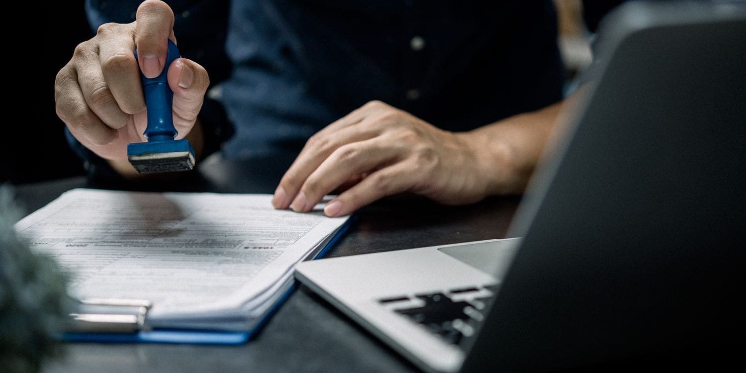 A close-up, eye-level shot of a person's hands at a dark desk, performing an official task. The person's right hand holds a blue self-inking stamp, hovering just above a document on a clipboard. The person is wearing a dark blue, long-sleeved collared shirt. Their left hand rests on the desk beside a silver laptop, which is angled towards them. The image has a shallow depth of field, keeping the hands, stamp, and document in sharp focus while the person's torso and the background are blurred. The overall mood suggests bureaucracy, approval, or immigration processing.